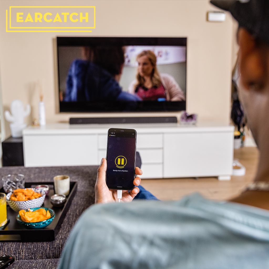 Een jonge man zit voor de televisie met in zijn linkerhand een telefoon waarop de Earcatch-app aan het afspelen is. In de linkerbovenhoek staat het gele logo van Earcatch.
