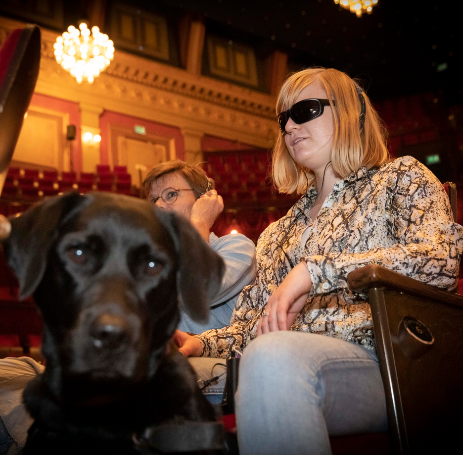 Een slechtziende vrouw zit in het theater met haar blindengeleidehond. Ze draagt een koptelefoon.